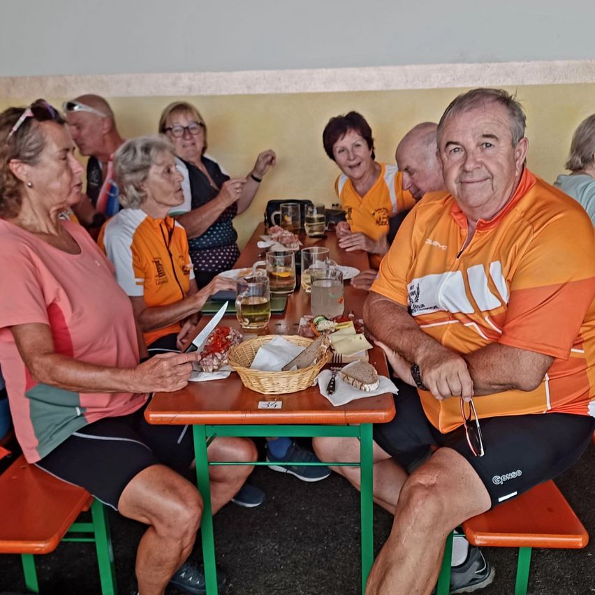A group of older adults, some in cycling jerseys, sit around a table with food and drinks, smiling and chatting.