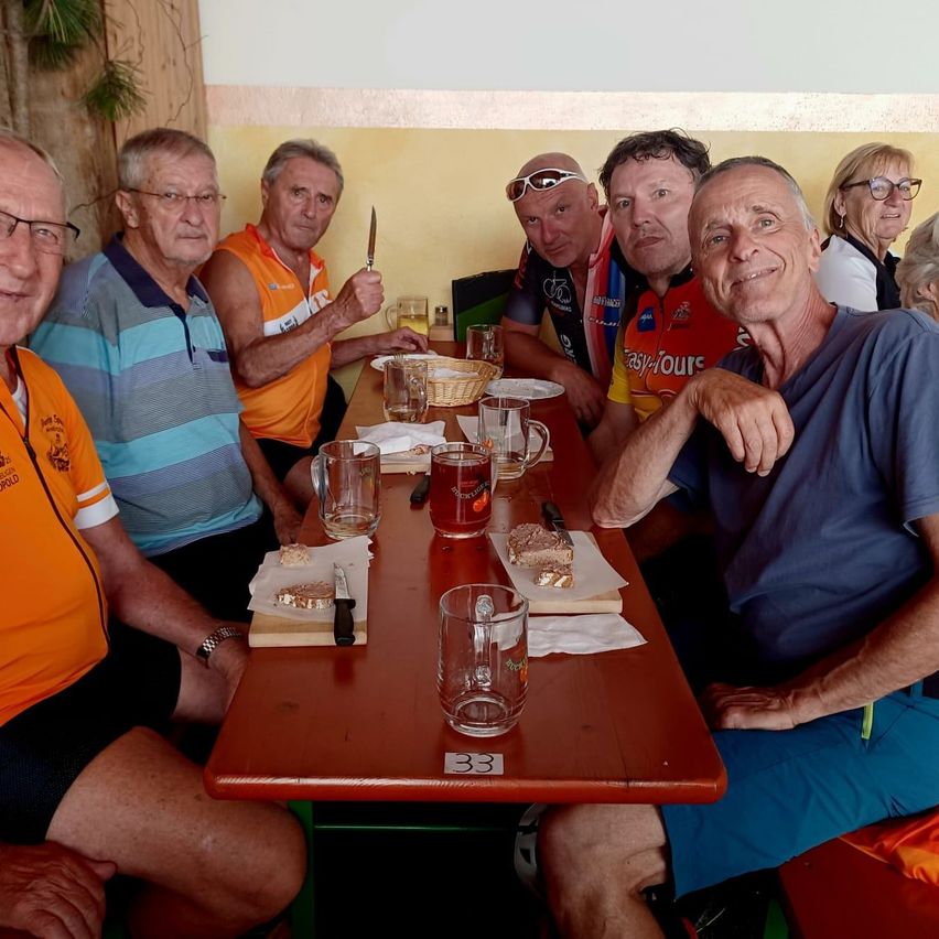 A group of older men in cycling jerseys and casual clothing are seated around a wooden table, smiling and posing for a photo. The table is set with glasses, plates, and a basket. One man holds a knife, and another has a drink.
