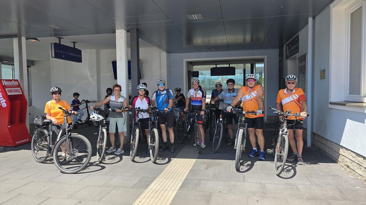 Radfahrer-Gruppe mit Helmen und Fahrrädern vor einem Bahnhofsgebäude in Österreich.
