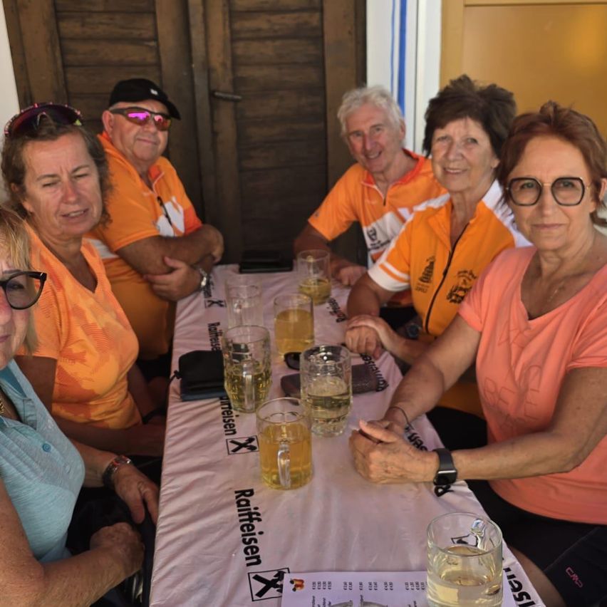 Eine Gruppe von Menschen in orangefarbenen Shirts sitzt um einen Tisch mit Biergläsern, lächelt und posiert für ein Foto.