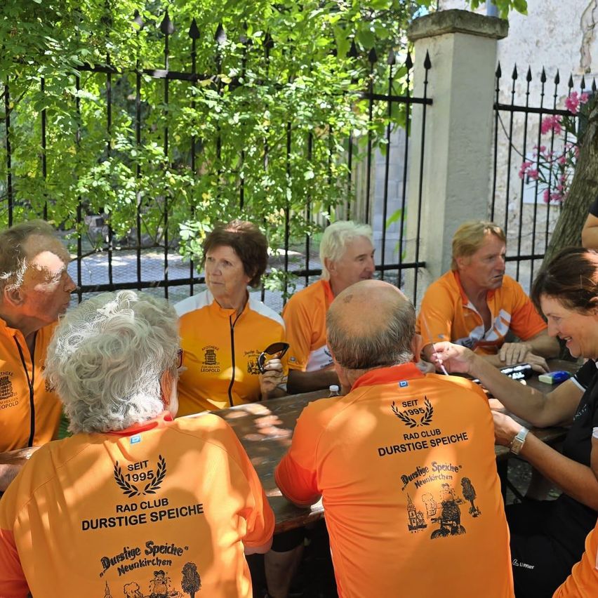 Eine Gruppe von Menschen in orangefarbenen T-Shirts mit 'Rad Club Durstige Speiche' bedruckt sitzt draußen um einen Tisch herum. Sie unterhalten sich.