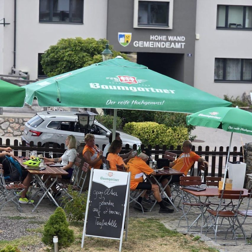 Eine Gruppe von Menschen sitzt unter grünen Sonnenschirmen an Tischen in einem Außenrestaurant. Ein Schild steht auf dem Boden, und ein Auto ist hinter dem Zaun geparkt.