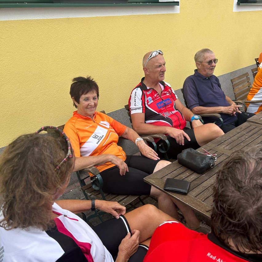 Eine Gruppe älterer Erwachsener in Fahrradtrikots sitzt um einen Holztisch herum, lächelt und plaudert. Auf dem Tisch liegen eine schwarze Tasche und ein schwarzer Geldbeutel. Die Wand hinter ihnen ist gelb.
