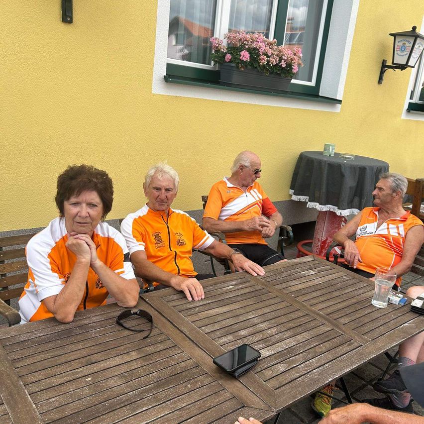 Vier ältere Erwachsene in orangefarbenen und weißen Radsporttrikots sitzen um einen Holztisch herum vor einem gelben Gebäude. Eine Frau mit kurzen Haaren und einer Brille auf dem Tisch sieht nachdenklich aus.