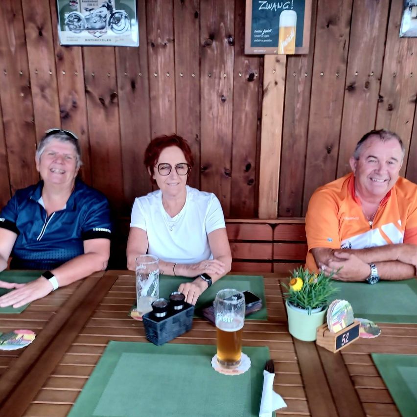 Drei Personen sitzen an einem Tisch mit Getränken und einer Pflanze. Die Frau in der Mitte trägt eine Brille. Hinter ihnen ist eine Holzwand mit zwei Schildern.