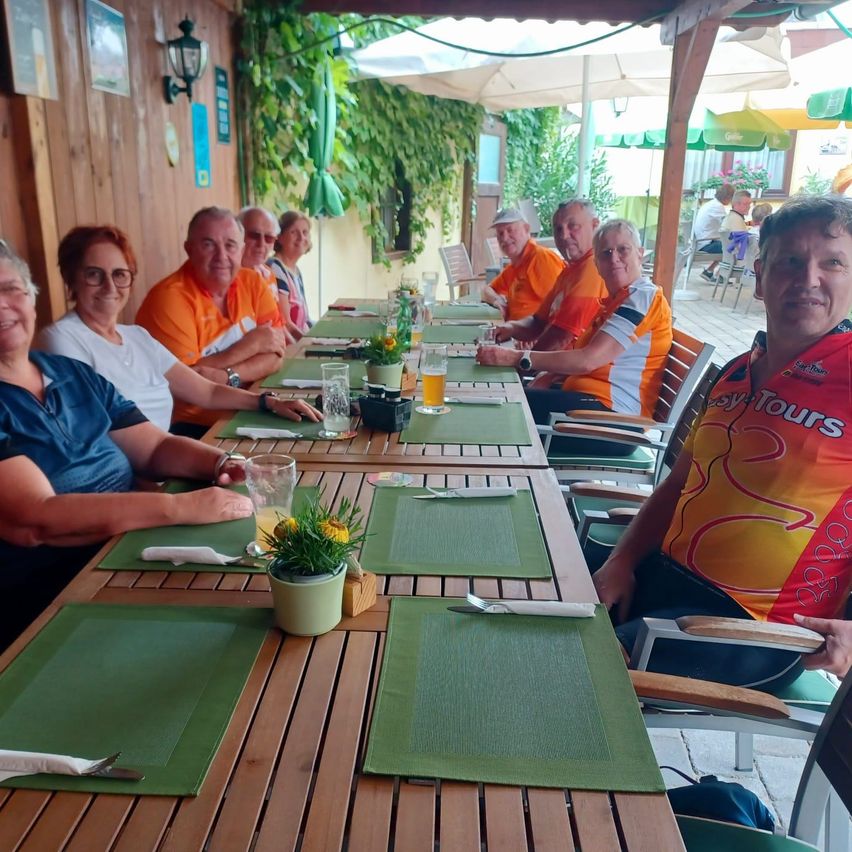 Eine Gruppe von Menschen in orangefarbenen und weißen Shirts sitzt an einem langen Tisch in einem Restaurant. Sie lächeln und posieren für ein Foto.