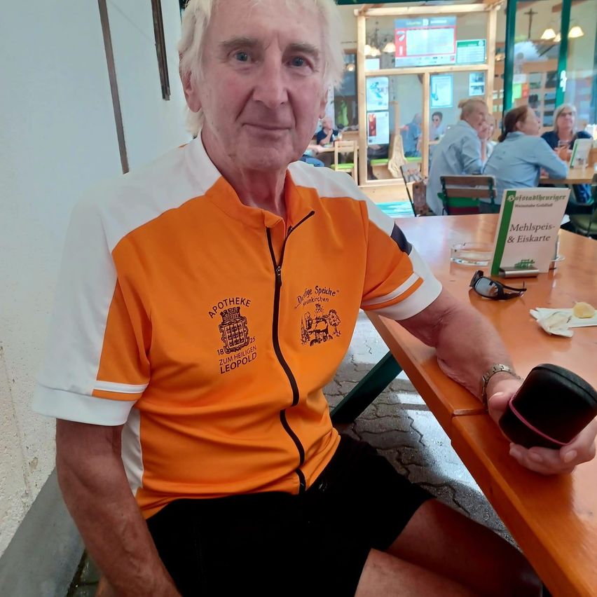 Ein älterer Mann in einem orange-weißen Radtrikot mit Logo und Text sitzt an einem Tisch in einem Restaurant.