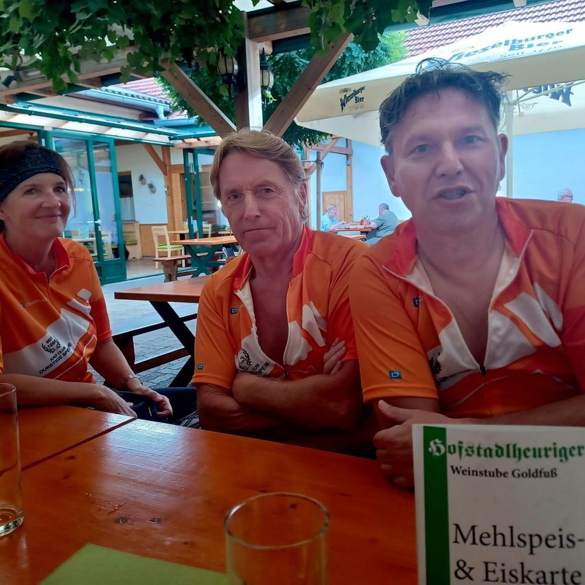 Drei Personen in orangefarbenen Shirts sitzen an einem Tisch in einem Restaurant, mit einem Menükarten vor ihnen. Der Mann auf der rechten Seite hat seinen Mund geöffnet.