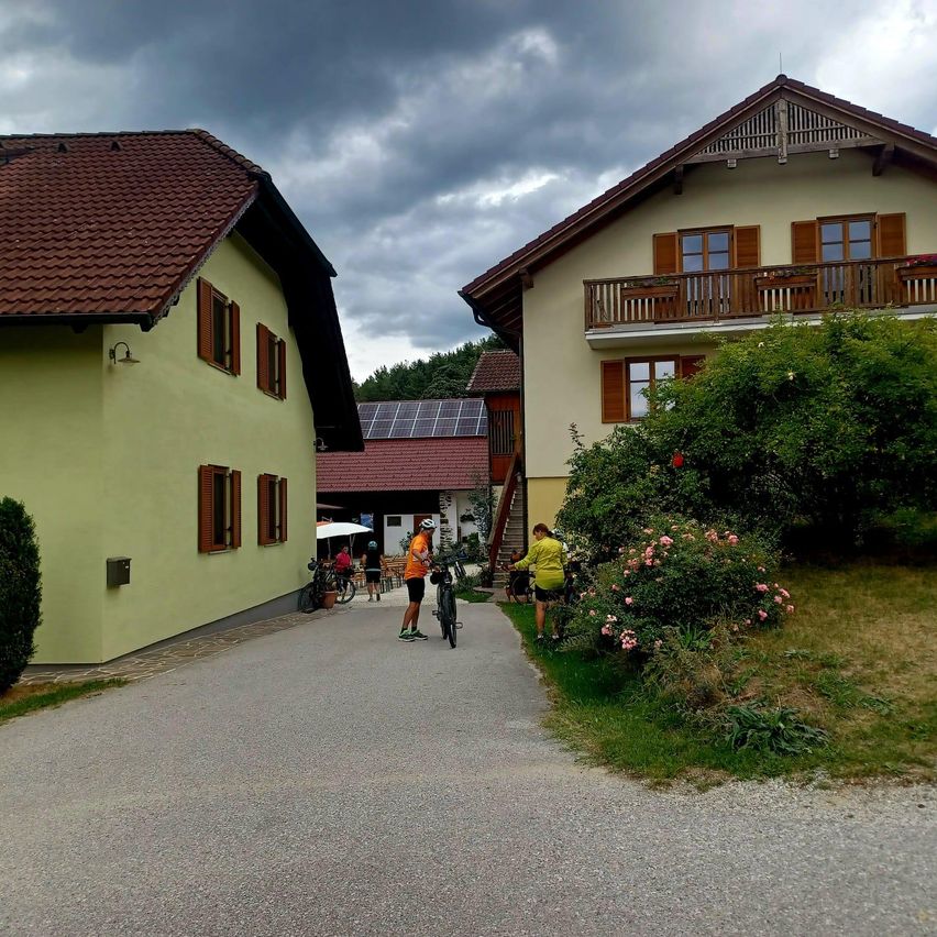 Zwei Personen stehen mit ihren Fahrrädern auf einer Einfahrt. Eine Person trägt einen Helm. Es gibt Häuser auf beiden Seiten der Einfahrt.