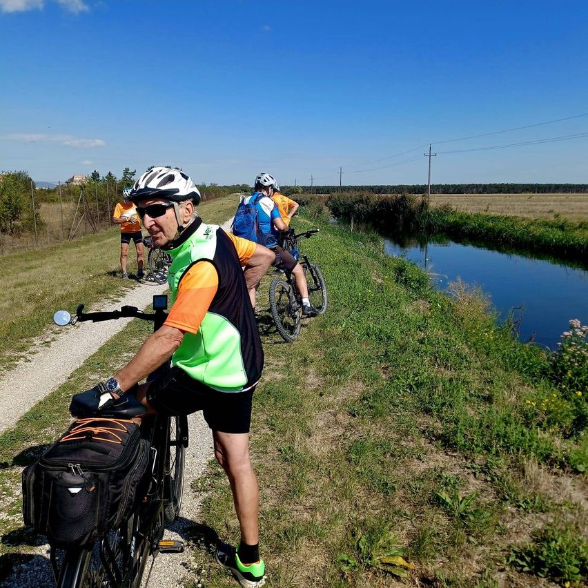 Eine Gruppe von Radfahrern fährt auf einem Pfad neben einem Kanal, wobei einer der Radfahrer eine reflektierende Weste trägt. Die Landschaft ist ländlich, mit einem klaren Himmel darüber.