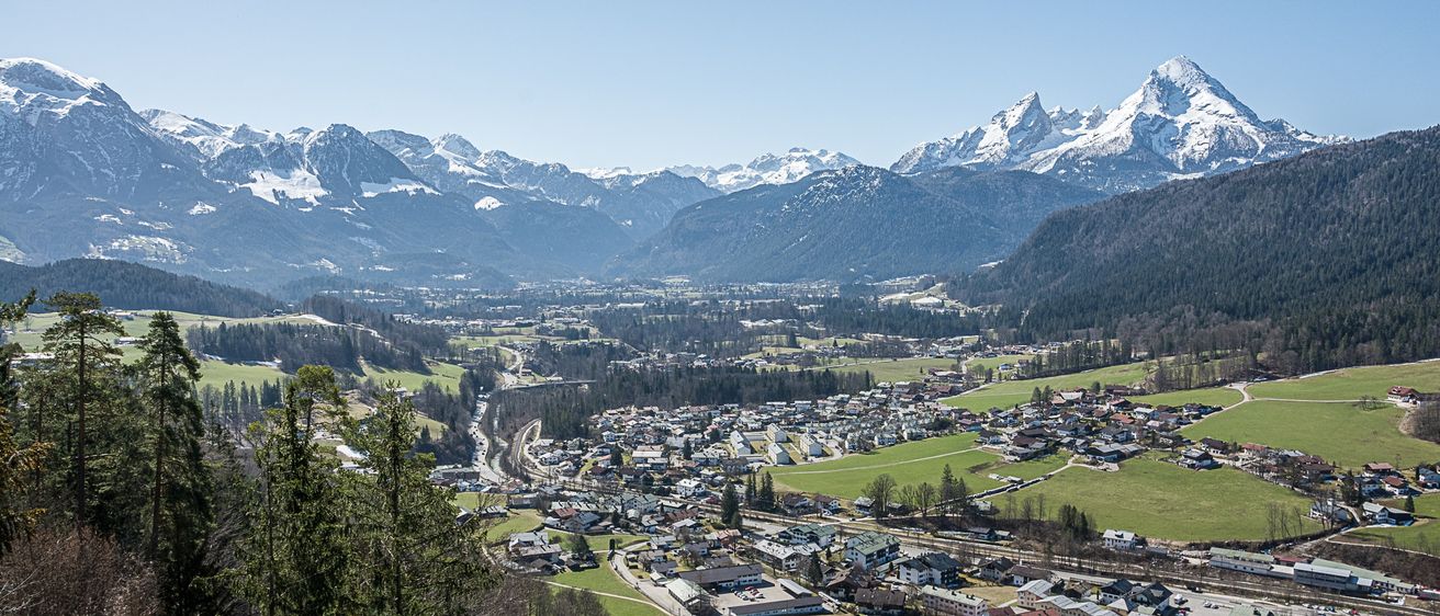 Luftaufnahme einer Stadt zwischen verschneiten Bergen und üppigen grünen Feldern unter einem klaren blauen Himmel.