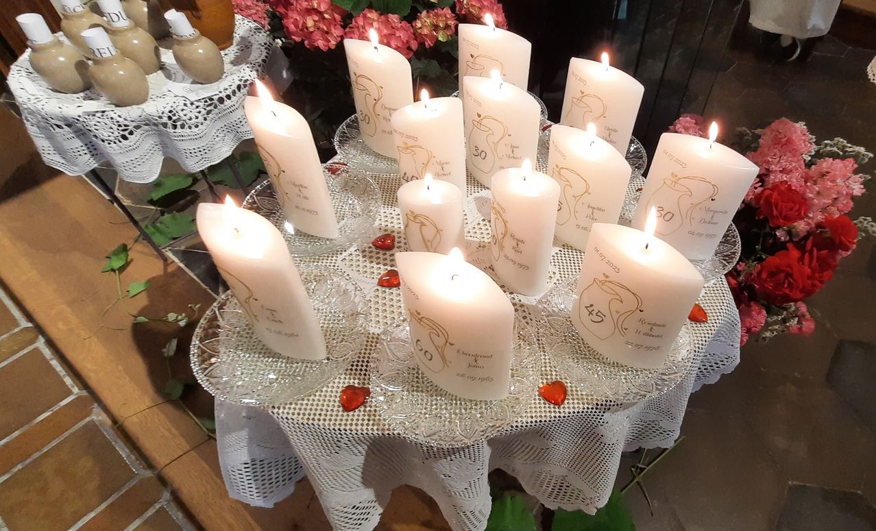 Ein Tisch hält mehrere brennende weiße Kerzen mit Altersinschriften darauf, jede Kerze in einem Glashalter, umgeben von rosa Blumen und Spitze.