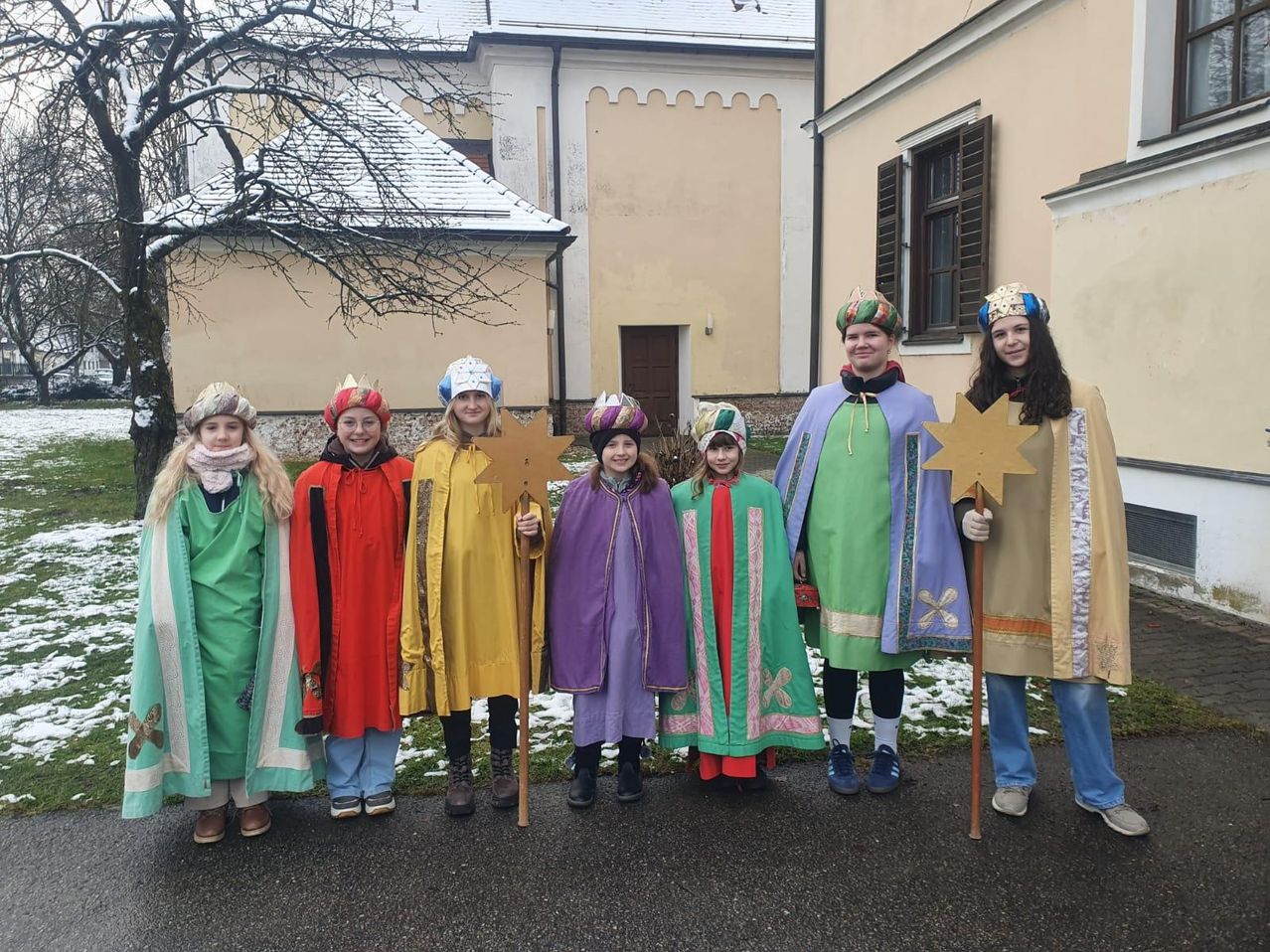 Sieben Kinder in farbenfrohen Kostümen und mit sternförmigen Requisiten posieren für ein Foto vor einem Gebäude mit Schnee auf dem Dach.