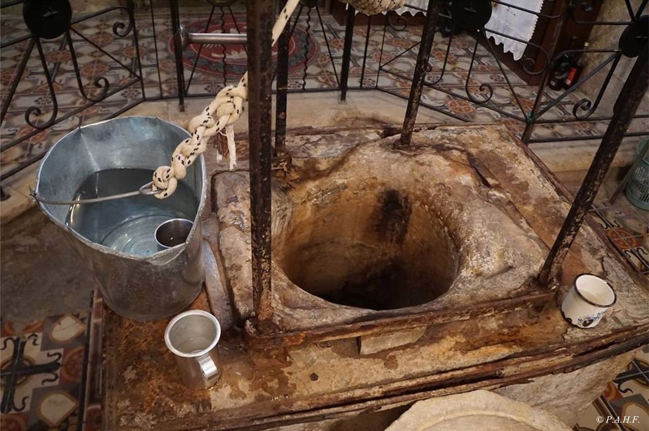 An old baptismal font with a bucket and a cup sits on a tiled floor with a rusted metal frame and a rope.