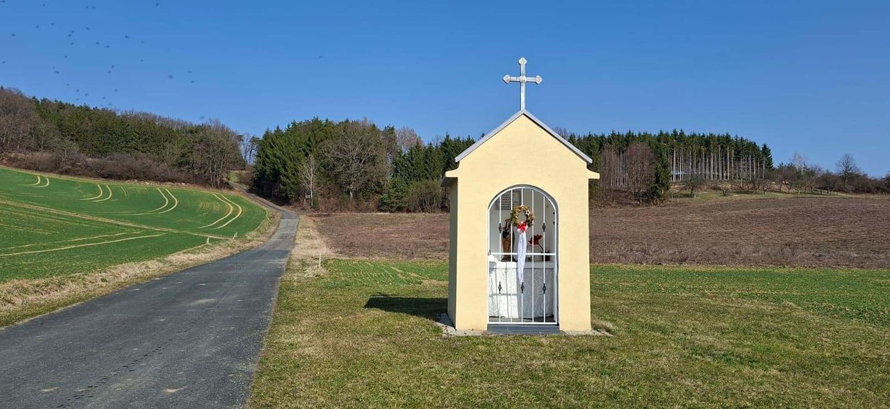 A small chapel with a cross on top is located on a grassy field, surrounded by trees and a dirt road.