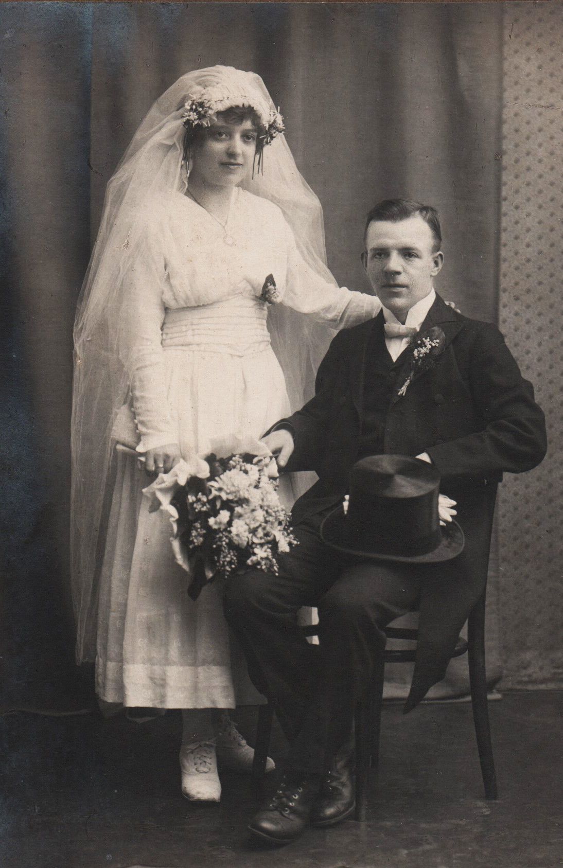 Ein altes Schwarz-Weiß-Foto eines frisch vermählten Paares. Die Braut trägt ein weißes Kleid und Schleier, während der Bräutigam in einem Anzug mit einer Blume an der Revers ist. Sie halten Händchen und sitzen zusammen.