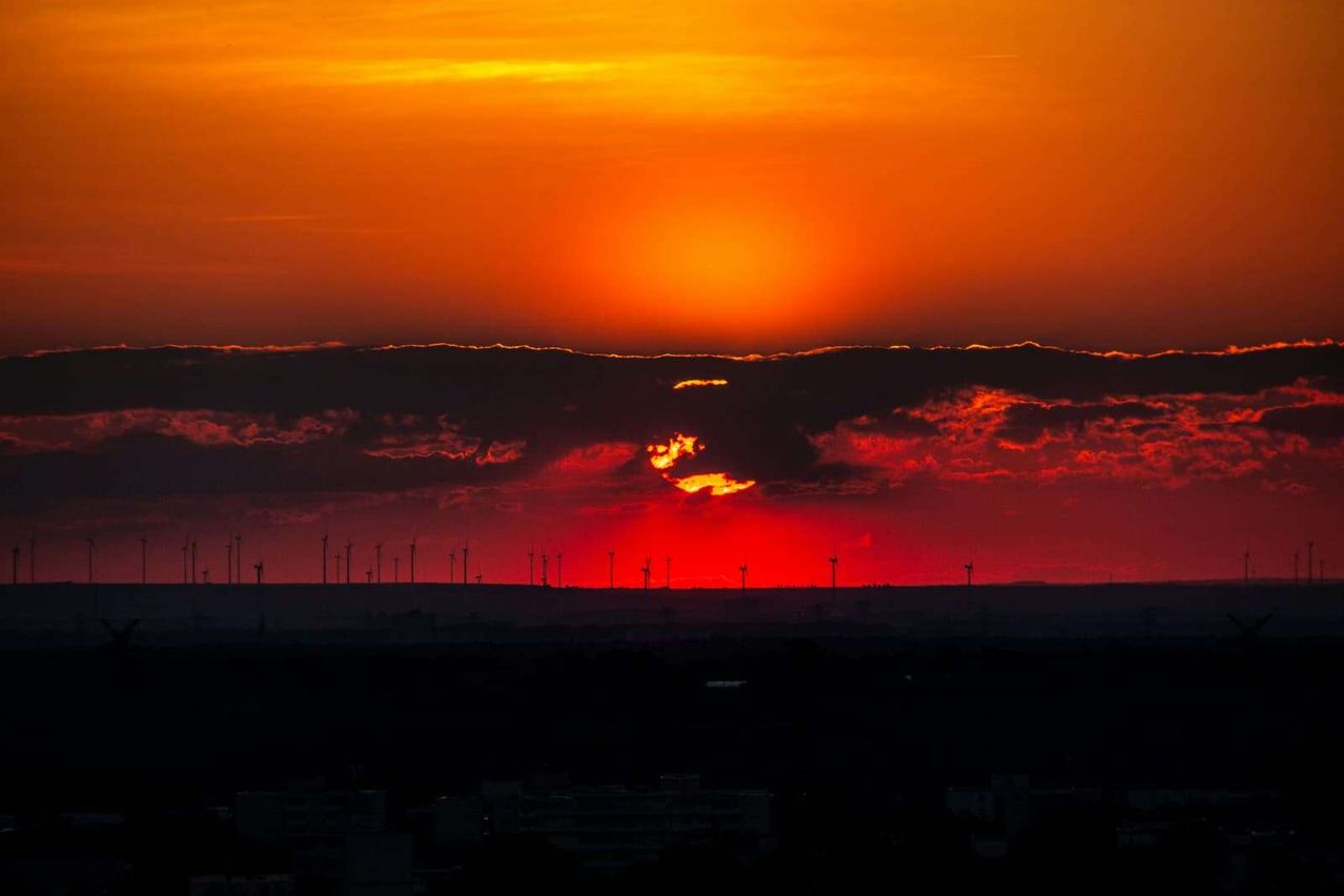 Ein Sonnenuntergang über einer Landschaft mit Silhouetten von Windturbinen. Der Himmel ist eine Mischung aus orange und roten Farbtönen.