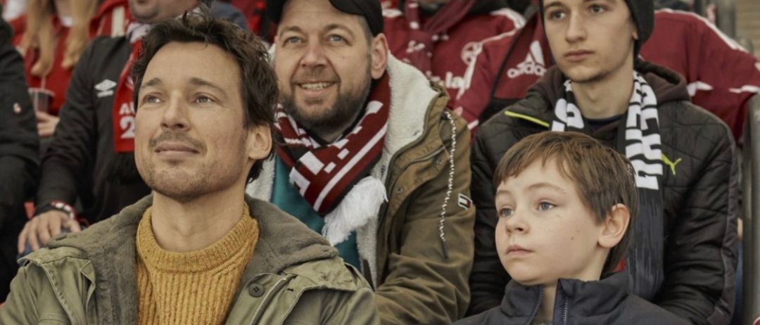 Eine Gruppe von Menschen sitzt in einem Stadion. Ein Mann mit Bart und Schal lächelt neben einem Jungen in einer blauen Jacke.