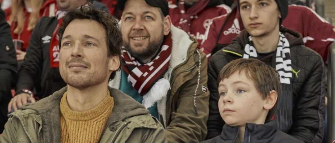 Zwei Erwachsene und zwei Kinder, von denen eines ein gelbes Sweatshirt und eine grüne Jacke trägt und das andere eine marineblaue Weste mit Logo, beobachten ein Spiel in den Rängen.