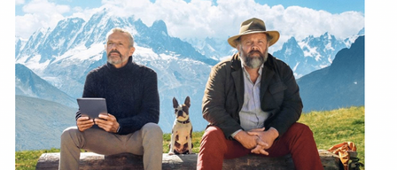 Two men sit on a log in a grassy field, one holding a tablet, the other with a hat and a small dog. The background features mountains and a clear blue sky.