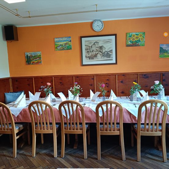 Ein Esszimmer mit Holzstühlen und -tischen, ein langer Tisch ist mit Blumen, Stoff und Besteck gedeckt. Es gibt Gemälde an der Wand und eine Uhr darüber. Der Raum hat Holzwände und einen gefliesten Boden.