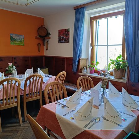 Ein Esszimmer mit Holztischen und Stühlen, mit Blumen, Servietten und Besteck gedeckt. Orange Wände und ein Fenster mit blauen Vorhängen. Topfpflanzen auf der Fensterbank.