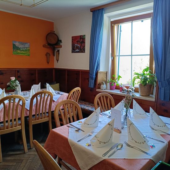 Ein Esszimmer mit Holztischen und Stühlen, mit Blumen, Servietten und Besteck gedeckt. Orange Wände und ein Fenster mit blauen Vorhängen. Topfpflanzen auf der Fensterbank.