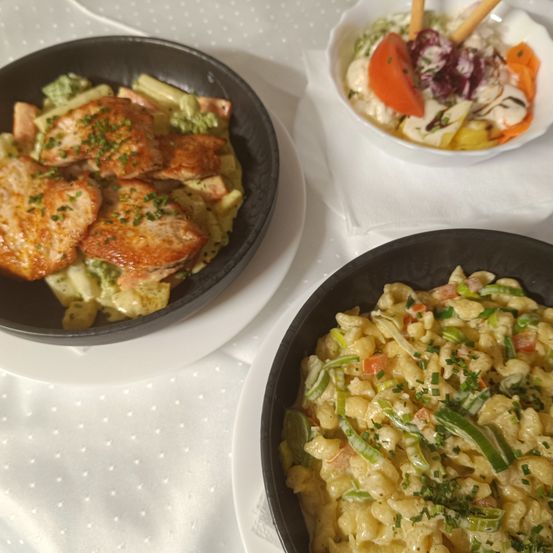 Zwei Teller mit Essen auf einer weißen Tischdecke, einer mit Pasta und Gemüse, der andere mit einem Fleischgericht und einem Salat in einer Schüssel.