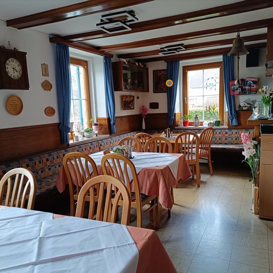 Ein leerer Essbereich mit Holzmöbeln, weißen Tischdecken und Blumendekorationen. Der Raum hat Holzwände, blaue Vorhänge und eine Holzdecke mit einer hängenden Glocke.