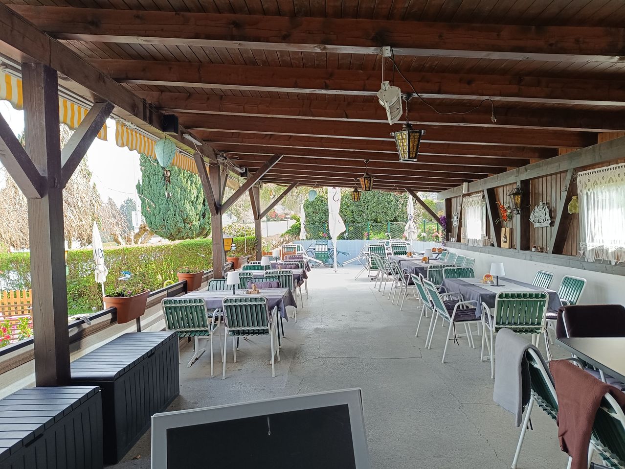 Ein Restaurant im Freien mit einer Terrasse. Es hat Holzbalken, Lampen und Stühle um die Tische herum. Es gibt auch Pflanzen und Büsche in der Umgebung.
