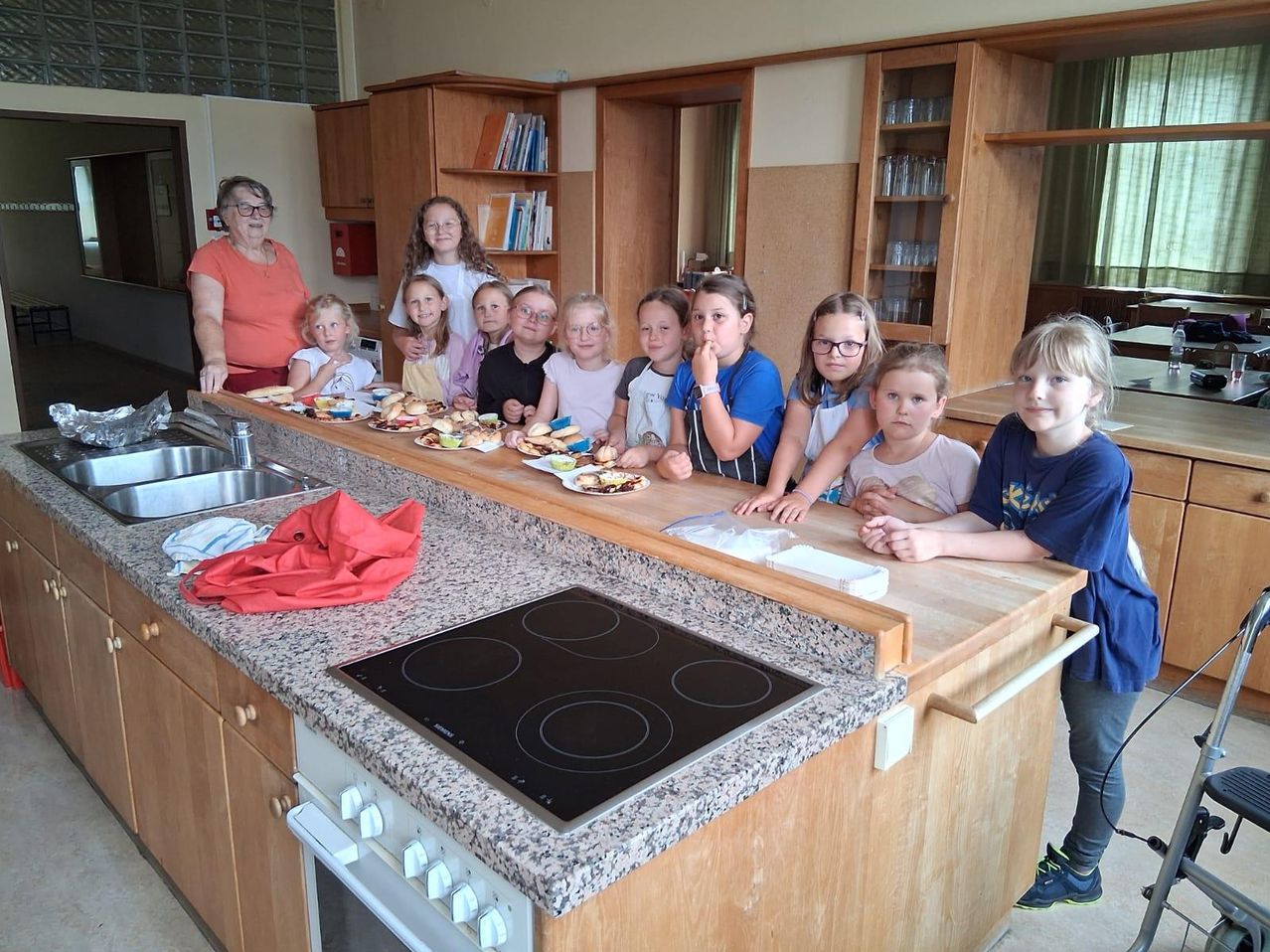 Eine Gruppe von Kindern und einer erwachsenen Frau befinden sich in einer Küche und genießen Essen von Tellern vor sich. Die Küche hat Holzschränke, ein Waschbecken und einen Herd.