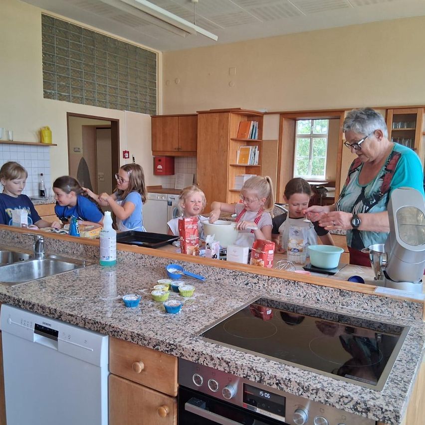 Eine ältere Frau steht an einer Küchentheke und leitet mehrere junge Kinder bei einer Kochaktivität an. Die Kinder tragen Schürzen und haben Schüsseln, Tassen und Zutaten vor sich. Ein Geschirrspüler und eine Spüle sind sichtbar.