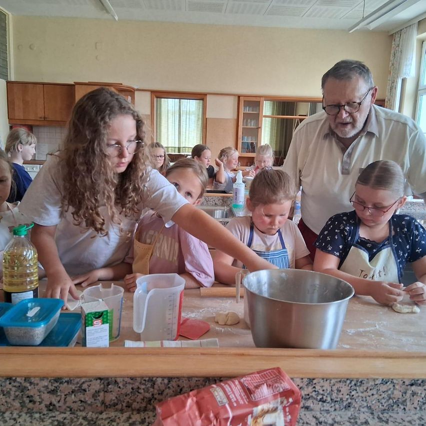 Eine Gruppe von Kindern und Erwachsenen bereitet in einer Küche Essen zu. Sie tragen Schürzen und arbeiten an einem Küchentresen mit verschiedenen Zutaten. Ein erwachsener Mann beaufsichtigt sie.