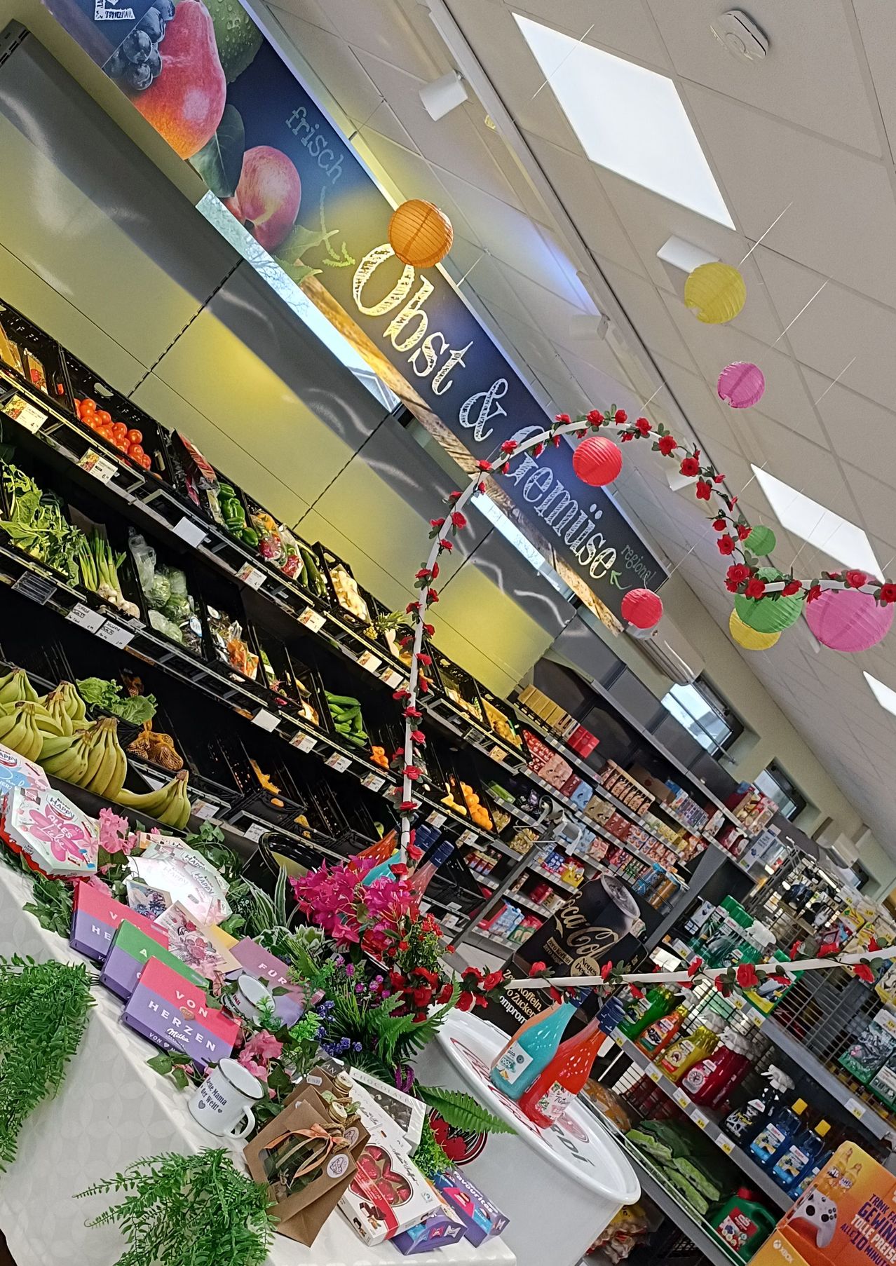A supermarket aisle with fresh produce on display, colorful flowers, and decorative paper lanterns hanging from the ceiling. The aisle features a banner with text in a foreign language.