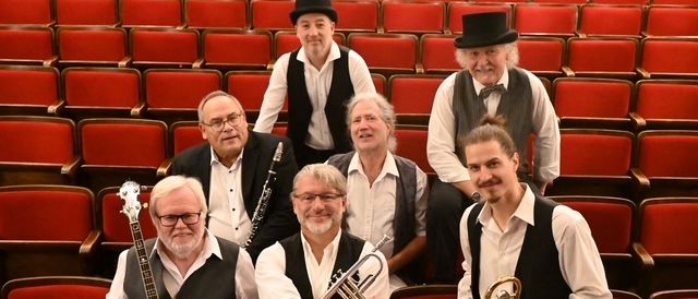 Eine Gruppe von Musikern in schwarzen Westen und weißen Hemden posiert in einem rotgesessenen Auditorium mit Musikinstrumenten.