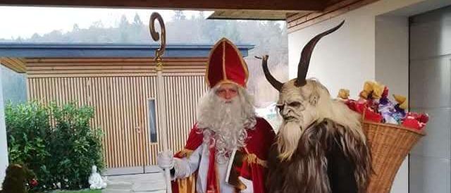 Zwei Personen in Santa Claus- und Krampus-Kostümen stehen in der Nähe eines Hauses. Der als Santa Claus gekleidete trägt einen Stab und ein Buch. Krampus trägt einen Korb voller Geschenke.