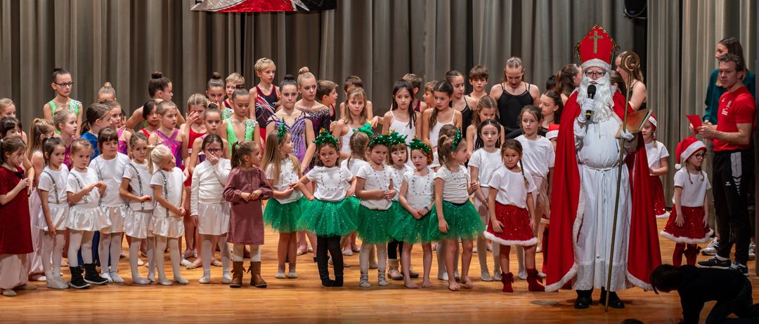 Eine Gruppe junger Kinder in verschiedenen Kostümen steht auf der Bühne mit dem Weihnachtsmann. Sie tragen unterschiedliche farbige Tüllröcke und Schuhe. Hinter ihnen hängt ein Banner von der Decke.
