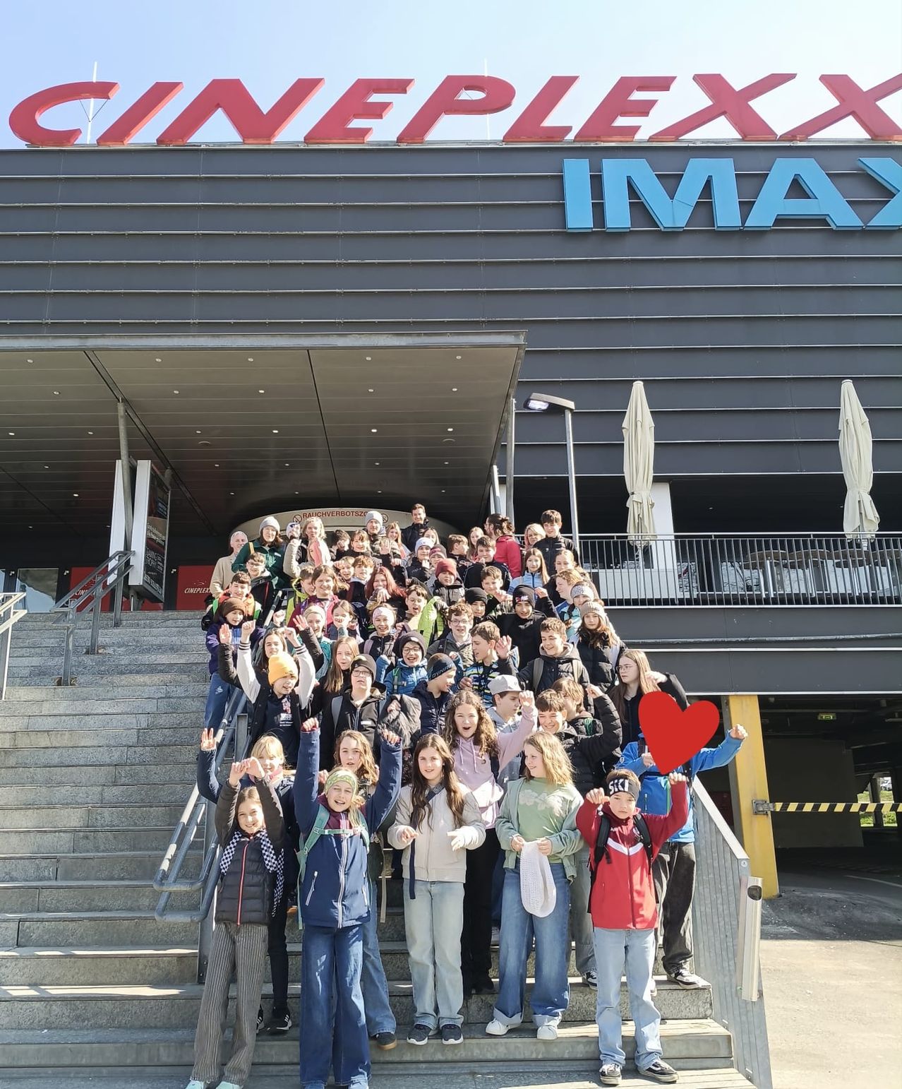 Eine Gruppe von Menschen, wahrscheinlich Schüler, posiert für ein Foto vor dem Cineplex IMAX-Kino. Sie stehen auf den Stufen, lächeln und winken, wobei der Name des Gebäudes deutlich über ihnen steht.