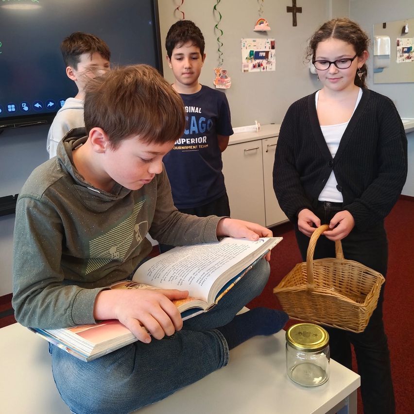 Ein Junge liest ein Buch, während er auf einem Tisch in einem Klassenzimmer sitzt, andere Kinder beobachten ihn.