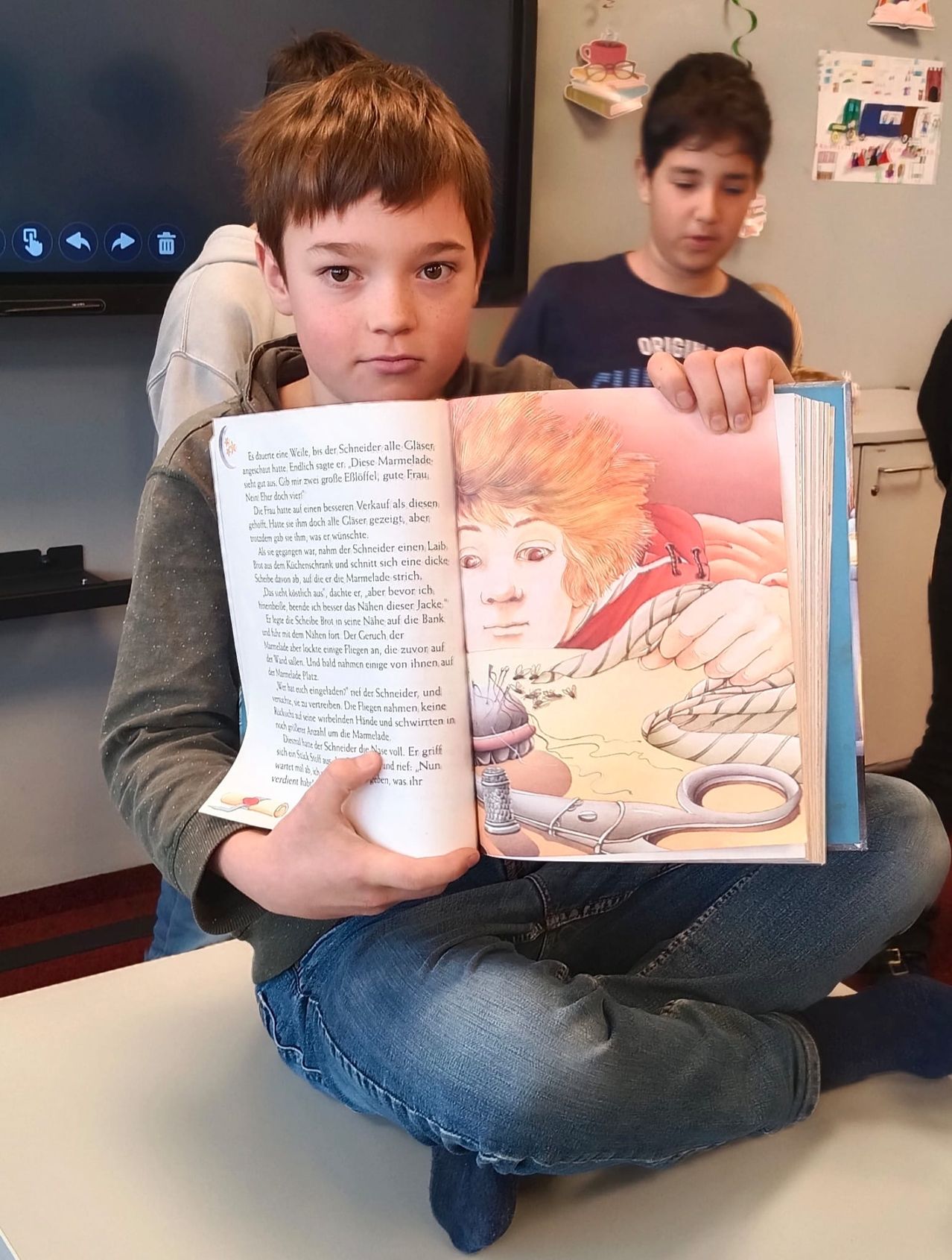 Ein Junge liest ein Buch mit einer Illustration eines Jungen mit roten Haaren, der Scheren hält. Ein anderer Junge hinter ihm schaut zu. Die Umgebung scheint ein Klassenzimmer zu sein.