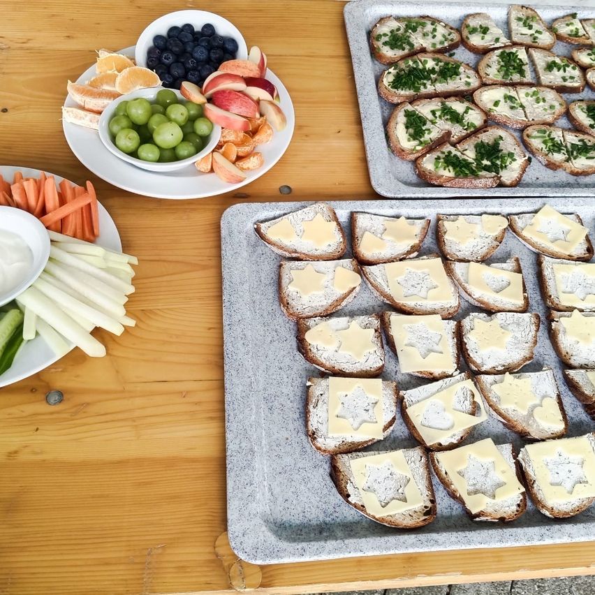 Auf einem Holztisch stehen mehrere Tabletts mit Essen: Obst, Gemüse und Käsebrot.