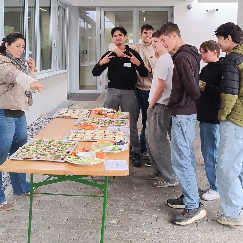 Eine Gruppe junger Männer und eine Frau versammelt sich um einen Tisch mit verschiedenen Speisen, wobei ein Mann zeigt. Sie scheinen sich in einem Innenraum zu befinden.