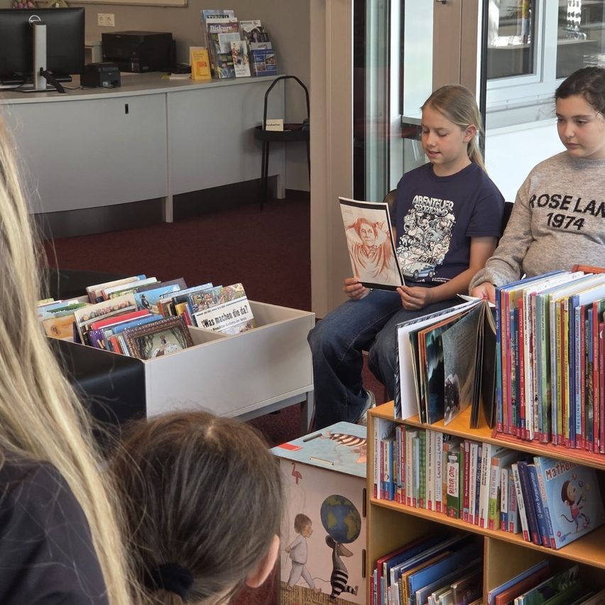 Zwei Mädchen sitzen in einer Bibliothek und lesen Bücher. Eine von ihnen hält ein Buch mit einer Zeichnung. Ein Regal voller Bücher steht vor ihnen.