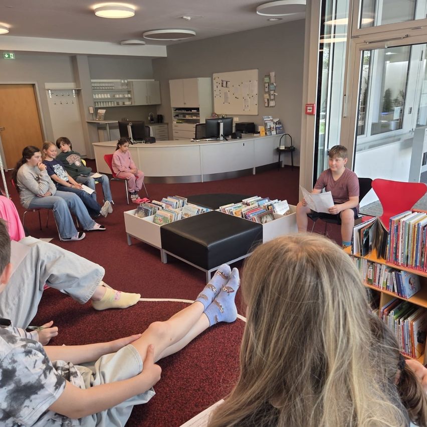 In einer Bibliothek sitzen eine Gruppe von Kindern um einen Tisch und hören einem Jungen zu, der laut vorliest. Ein anderer Junge sitzt auf einem Stuhl und liest ein Buch.