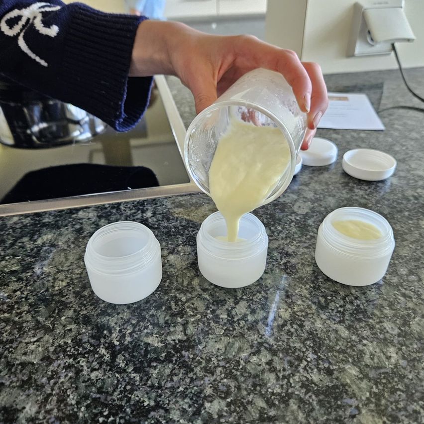 Eine Hand gießt eine Flüssigkeit aus einem Messbecher in einen Behälter auf einem Marmortisch, mit drei anderen Behältern davor.