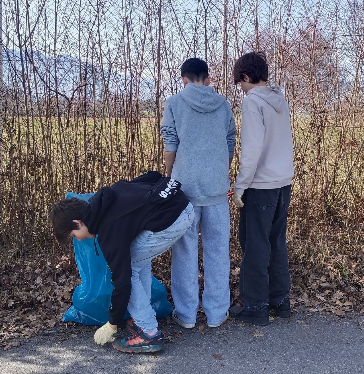 Three boys in hoodies and pants clean up trash near a bush. One boy is picking up litter while the others watch.