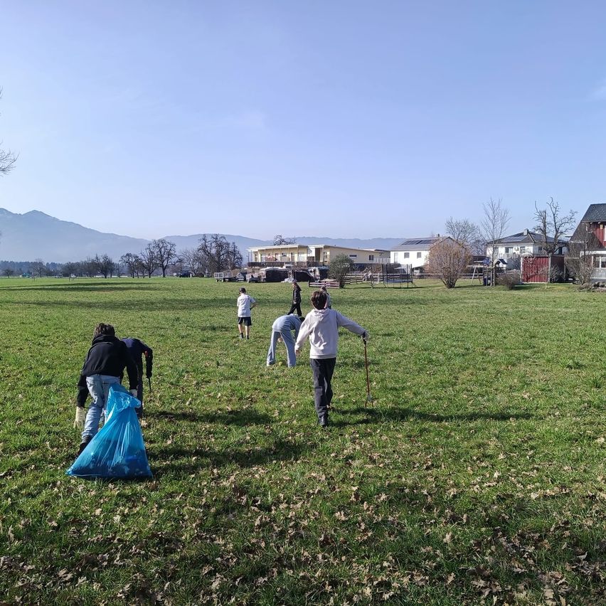 A group of people in a field are picking up trash with bags. Some are holding sticks. In the distance, there are houses and mountains.