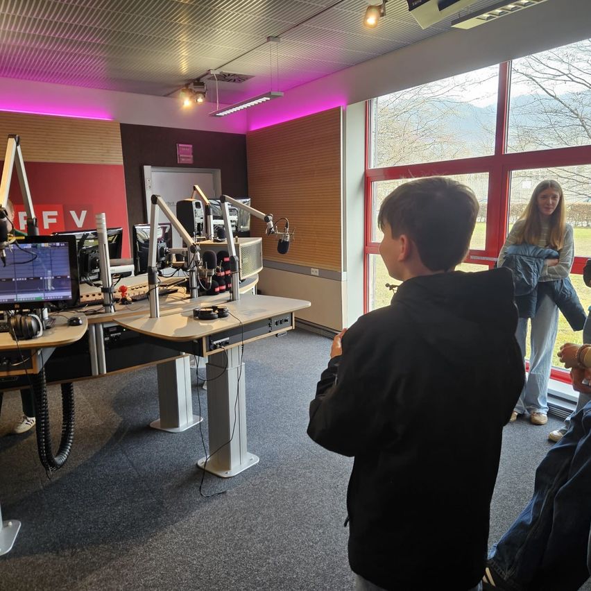 A boy looks into a radio studio with a desk full of equipment. A woman stands beside a glass wall with a view of the outside.