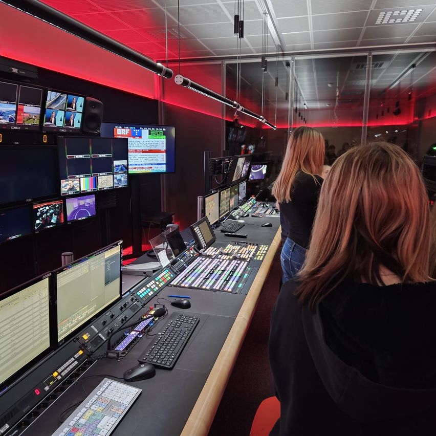 A studio control room with multiple monitors, keyboards, and a large screen. Two women observe the screens from behind the control desk.