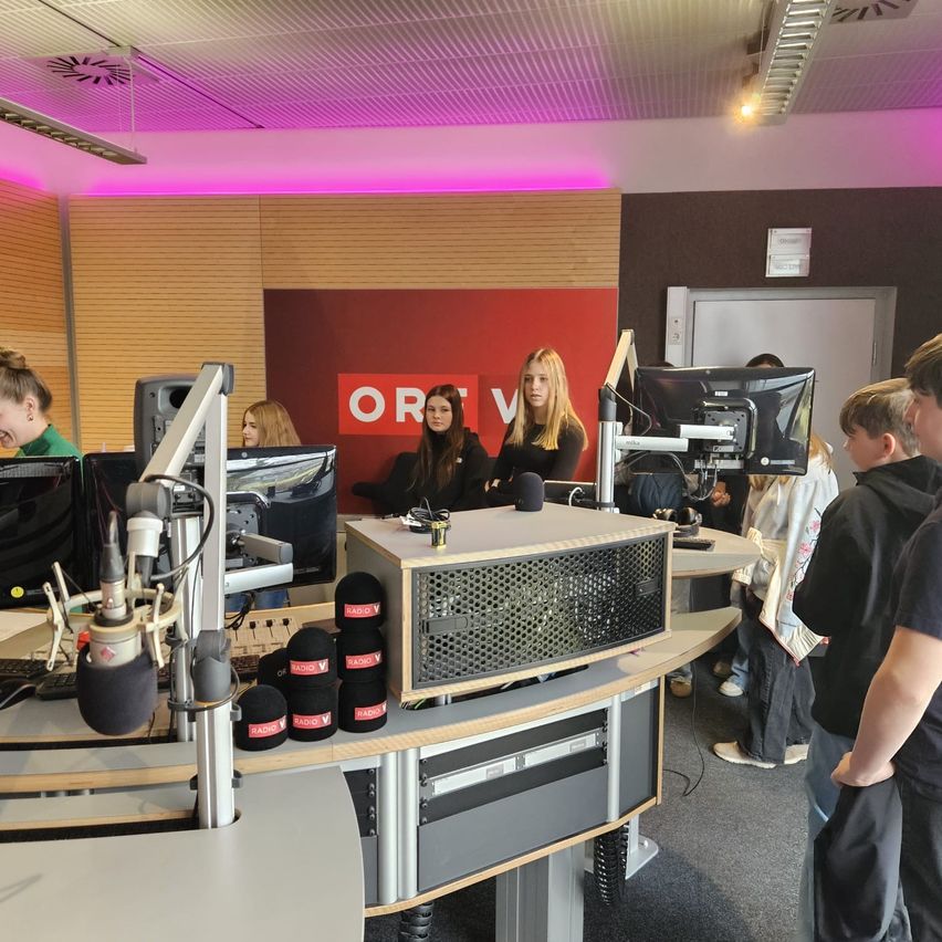 A group of people in a radio studio with a red ORV sign. Four women sit at a table with monitors and microphones. People stand around them.